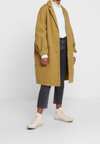 Abrigo largo mostaza, de corte holgado, abotonado, sobre una camiseta azul clara. Combinado con jeans grises y zapatillas altas beige.