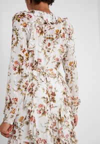 Robe florale blanche avec des détails froncés, des manches longues et une taille à nouer. Le tissu a une texture transparente avec des accents floraux métalliques.