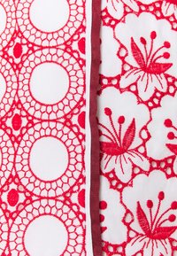 Tissu brodé présentant des motifs floraux rouges et de dentelle circulaire sur un fond blanc, avec un détail de ruban bordeaux le long de la couture.