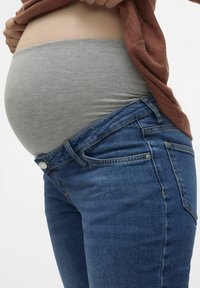 Těhotenské džíny v tmavě modrém denimu s pružným šedým pasem, střih obepínající postavu a světlé vyblednutí na přední straně; nošeny pod břichem.