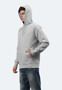 Felpa grigia con tasca frontale, cappuccio con cordoncino, polsini e orlo a coste. Indossata con jeans blu, che mostrano un design casual e una vestibilità comoda.