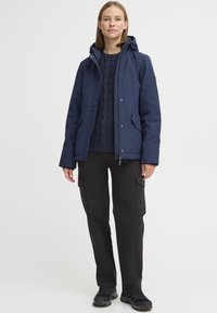 Veste imperméable navy avec capuche, fermeture éclair à l'avant et boutons-pression, associée à un pull en maille navy texturé et un pantalon cargo noir.