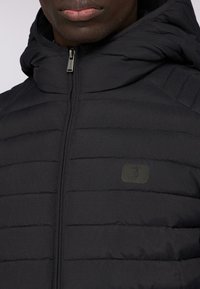 Svart pufferjacka med horisontellt quiltat mönster, hög krage och blixtlås. Har ett litet, diskret logopatch på bröstet.