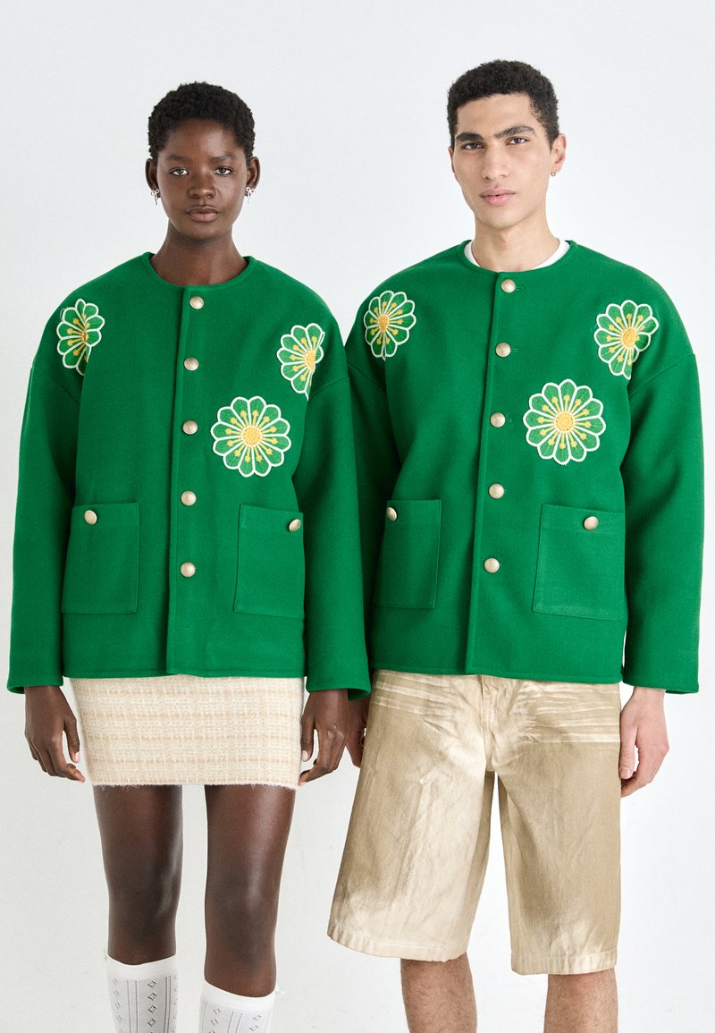 Grønne ullblandede jakker har store blomsterbroderier og gullknapper. Den mannlige modellen har på seg beige shorts; den kvinnelige modellen har et mønstret miniskjørt.