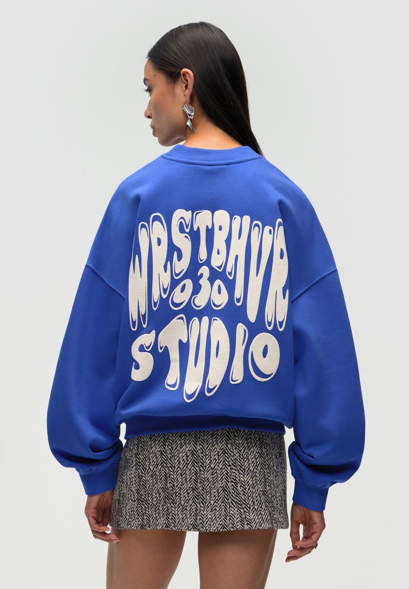 Sudadera azul de gran tamaño con un atrevido texto gráfico blanco en la espalda, escote redondo y mangas abullonadas. Combinada con una falda negra texturizada.