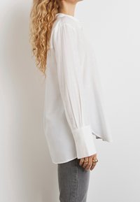 White long-sleeve blouse with button detail, relaxed fit, soft textured fabric, and wide cuffs, paired with gray denim jeans.
