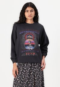 Felpa nera oversize con stampa grafica multicolore che rappresenta un sole e un paesaggio. Maniche lunghe e polsini a costine. Abbinata a una gonna floreale.