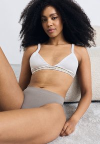 Hvid bralette med en Calvin Klein-logo bånd og højtaljede grå trusser på en model, der sidder imod et blødt tæppe. Krøllet mørkt hår.