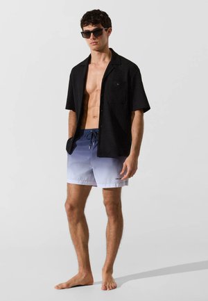 Giovane uomo con camicia nera a maniche corte aperta, pantaloncini da bagno blu a gradiente e occhiali da sole scuri, in piedi a piedi nudi su sfondo chiaro.