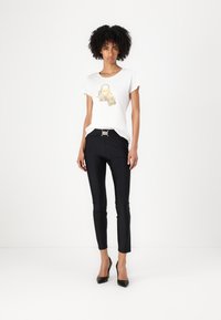 T-shirt bianca con design di lucchetto dorato; pantaloni neri slim-fit con cintura decorativa; il modello indossa scarpe col tacco nere.