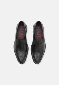 Mocasini negri din piele cu finisaj neted, având un model subtil cu logo și un interior căptușit roșu cu literele "HUGO".