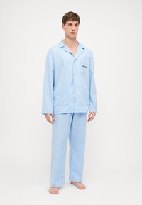 Lyseblå pyjamas-sett, laget av mykt stoff, med en knappedesign på toppen som har to frontlommer og matchende bukser, designet for komfort.