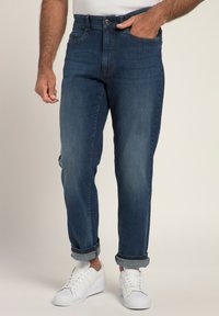 Jeans in denim blu scuro con taglio diritto, risvoltati alla caviglia, dotati di cinque tasche e chiusura con bottone. Indossati con sneakers bianche.