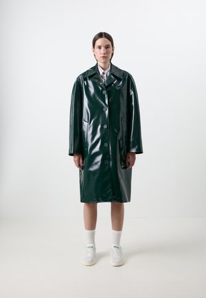 Manteau en vinyle vert et brillant avec un col à revers, des boutons sur le devant et des poches latérales. Le modèle porte des baskets blanches et des chaussettes, se tenant devant un fond uni.