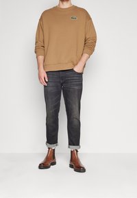 Hombre con un sudadera color beige con un logo de cocodrilo verde, jeans oscuros remangados y botines de cuero marrón, con la mano en el bolsillo.