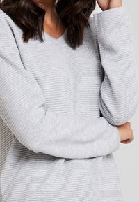 Vaaleanharmaa, teksturoitu neulepaita, jossa ribbineulos, V-aukko ja lasketut olkapäät. Pehmeää neulosta, pitkät hihat ja rento istuvuus.