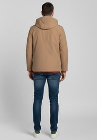 Chaqueta con capucha color marrón claro, de acabado liso, con un forro interior naranja en contraste. Llevada sobre jeans azules y zapatillas blancas con detalles verdes.