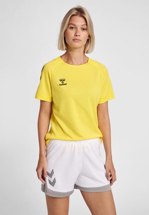 Maglietta sportiva gialla con finitura testurizzata, logo nero sul petto, abbinata a pantaloni corti bianchi con accenti grigi e dettagli a motivo.