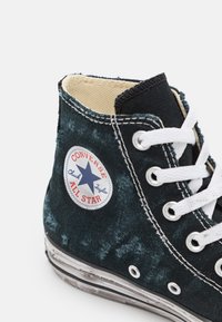 Zapatilla alta de lona negra con cordones blancos, suela de goma y parche del logo de Converse All Star con una estrella azul. Detalles de tejido texturizado y costuras.