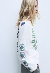 Blonde persoon die een wit longsleeve-shirt draagt met groene en blauwe 'Red Hot Chili Peppers'-logo's op de mouwen en de borst, staande in profiel.