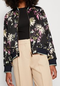 Bloemige bomberjack in zwart, met roze en groene patronen. Gouden rits, geribde navy mouwen en muntgroene accenten. Gecombineerd met beige broeken.