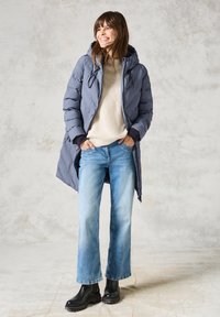 Vadderad grå kappa med huva, beige tröja, ljusblå utsvängda jeans och svarta ankelskotas. Slät yta och avspänt design.