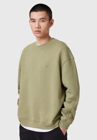 Sudadera verde oliva de tejido suave con cuello redondo, hombros caídos y un pequeño logo en relieve en el pecho. Presenta puños y dobladillo de canalé.