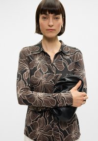 Ilga piedurkne melna blūze ar bežām ziedu rakstiem, ar apkakli un pogu aizdari. Modelis tur melnu, teksturētu sajūga somu.