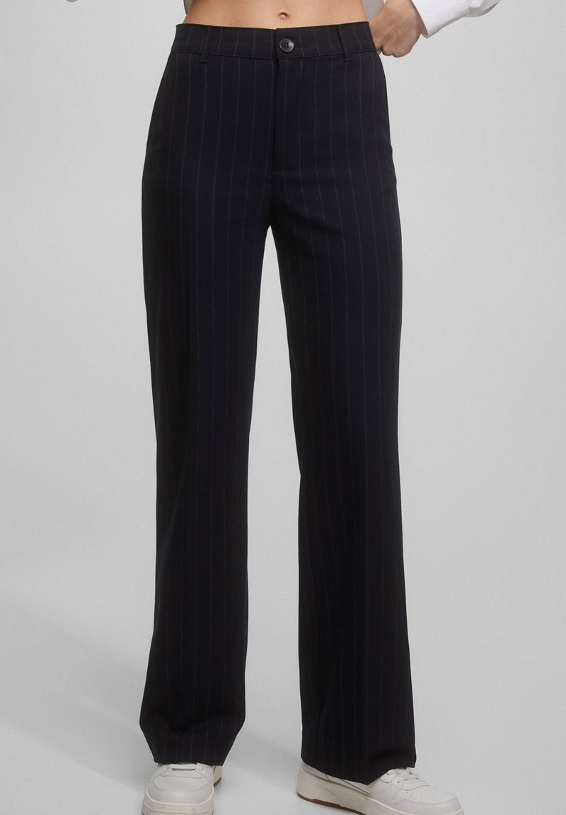 Pantalon large à fines rayures noires porté avec des baskets blanches et une chemise blanche rentrée, visible de la taille aux pieds.