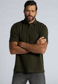 Polo vert olive avec une texture douce, patte de boutonnage à trois boutons, manches courtes et un logo discret sur la poitrine. Associé à un jean foncé.
