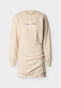 Μπεζ φόρεμα από φλις με μακριά μανίκια, στρογγυλή λαιμόκοψη, πιέτες στα πλαϊνά και λογότυπο "Calvin Klein" στο μπροστινό μέρος σε κόκκινο και λευκό. Απαλή υφή υφάσματος.