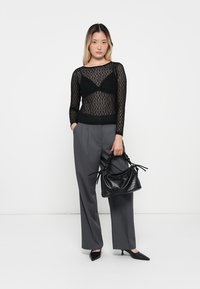 Schwarzes Spitzenoberteil mit langen Ärmeln und transparentem Design, kombiniert mit grauen, maßgeschneiderten Hosen. Accessoires sind eine kleine schwarze, strukturierte Handtasche.
