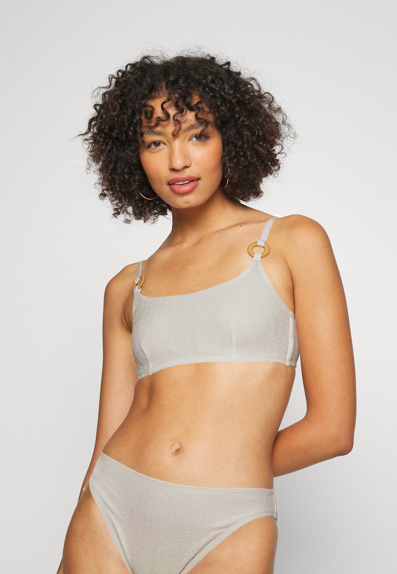 Conjunto de lingerie cinza claro, apresentando um sutiã texturizado com alças finas e acessórios circulares em tom dourado, acompanhado por um fundo de biquíni combinando.