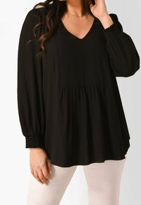 Svart långärmad topp med v-ringning, texturerad tyg, samlad midja och smockade manschetter, kombinerad med ljusfärgade byxor.