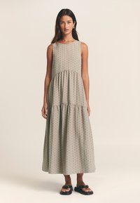 Maksi obleka iz mehkega, svetlo bež blaga z večnim krilom, brezrokavnim dizajnom in vzorcem majhnih črnih pik po celotni površini. Sandali z odprtimi prsti.