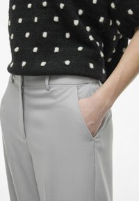 Pantalon gris ajusté avec une texture lisse et une fermeture éclair, associé à un pull noir à pois blancs. Détail de la main dans la poche.