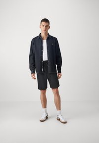 Cămașă de denim închis cu nasturi, maiou alb, pantaloni scurți negri, adidași albi cu accente negre, stând pe un fundal simplu.
