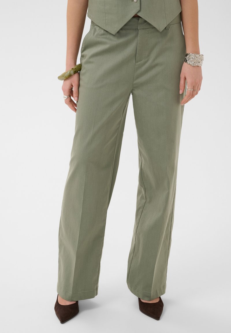 Femme portant un pantalon large vert olive, un gilet sans manches, des chaussures marron pointues, un bracelet vert au poignet gauche et un bracelet argenté au poignet droit.