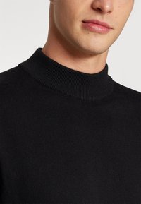 Suéter de punto negro con cuello alto, de textura suave y con canalé, que presenta un cuello ajustado y un acabado limpio alrededor del escote.