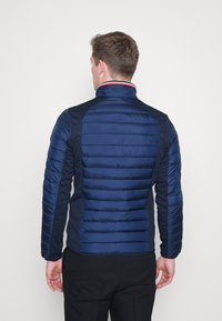 Mann trägt eine marineblaue gesteppte Jacke mit horizontaler Steppung, steht mit dem Rücken zur Kamera vor einem schlichten weißen Hintergrund.