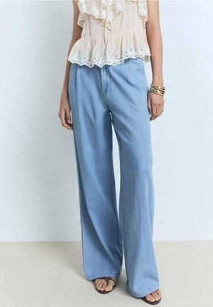 Person iført vide lyseblå jeans, en ærmeløs flæsebluse i cremefarve, sandaler og guldarmbånd, står mod en ensfarvet baggrund.