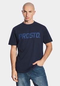 Granatowa bawełniana koszulka z krótkim rękawem z wyrazistym logo "PROSTO." w jaśniejszym odcieniu niebieskiego. Gładka tekstura, okrągły dekolt, luźny krój.