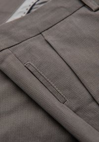 Gros plan sur un pantalon en tissu marron texturé montrant une poche avant ajustée et des détails de couture à la ceinture.