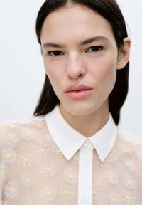 Blusa trasparente con motivo floreale, caratterizzata da un colletto e bottoni bianchi, con delicati accenti gialli su un tessuto leggero e trasparente.