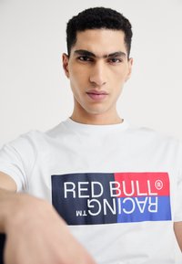 Balts kokvilnas T-krekls ar izteiktu grafisku logotipu: "RED BULL RACING" lieliem burtiem uz sarkanas un zilas taisnstūra fona.