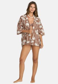 Kimono marrone a maniche corte con motivo bianco a foglie di palma; tessuto leggero. Scollo aperto con bottoni; vestibilità comoda. Indossato con un bikini sotto.