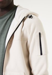 Veste à capuche en mélange de coton couleur écru, avec une finition texturée, des accents de fermeture éclair noirs et une poche zippée sur le côté de la manche.
