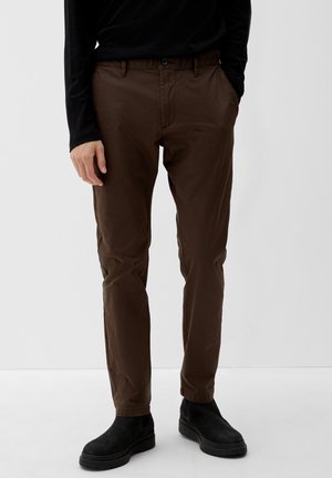 Mężczyzna ubrany w brązowe spodnie slim-fit, czarną koszulę z długim rękawem i czarne zamszowe buty, stojący na tle jednolitego tła.