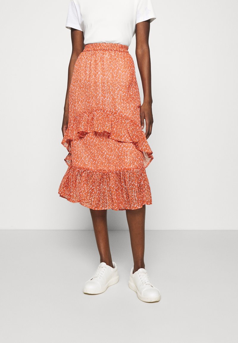 Femme portant un t-shirt blanc et une jupe midi orange à motif floral avec des détails volantés, associée à des baskets blanches, debout sur un sol uni.