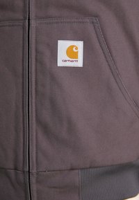 Dark gray jacket with a zipper, made of sturdy fabric. Features a white label with an orange logo. Includes a front pocket with a ribbed hem.